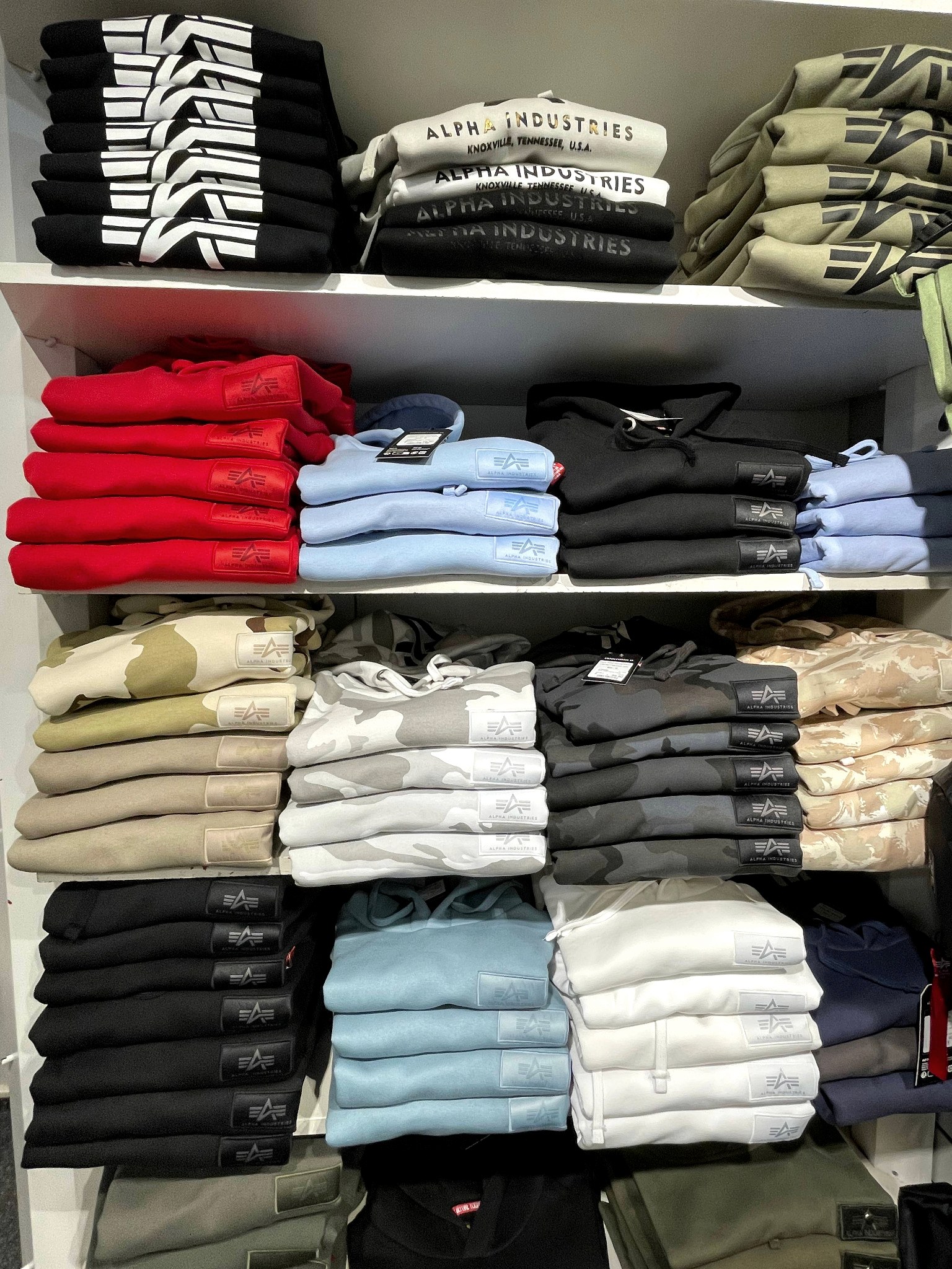 Regal mit gestapelten Alpha-Industries-Hoodies und Sweatshirts in Rot, Hellblau, Camouflage und Schwarz